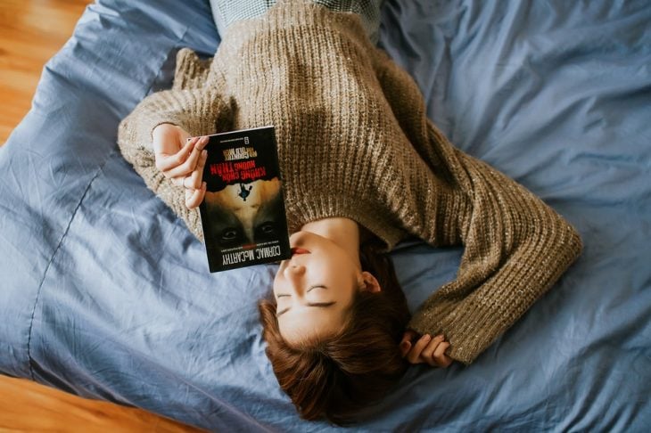 Cica recostada en una cama con sabanas azules, leyendo un libro de Stephen King, usando suéter café y acariciando su cabello rojizo