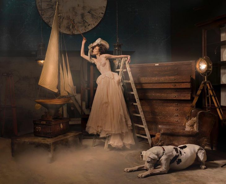 Vestidos de novia vintage confeccionados por la diseñadora Joanne Fleming, sesión de fotos anitguas, mujer parada en escaleras, con vestido al lado de un perro gran danés blanco con manchas negras y un barco de madera a escala