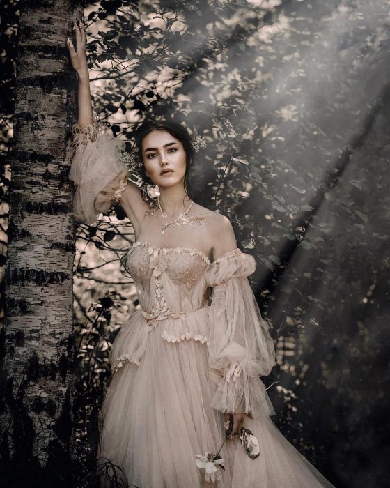 Vestidos de novia vintage confeccionados por la diseñadora Joanne Fleming, retrato de mujer al lado de un árbol con la luz del atardecer, usando vestido de tul vaporoso de color rosa pálido, sosteniendo una flor en la mano