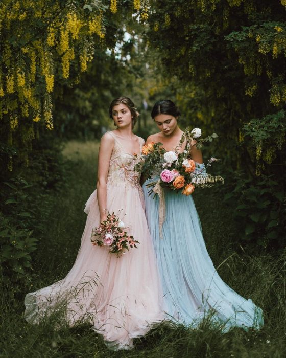 Vestidos de novia vintage confeccionados por la diseñadora Joanne Fleming, dos mujeres en medio del campo con vestidos elegantes vintage color rosa y azul de tela tul, sosteniendo un ramo de rosas en las manos