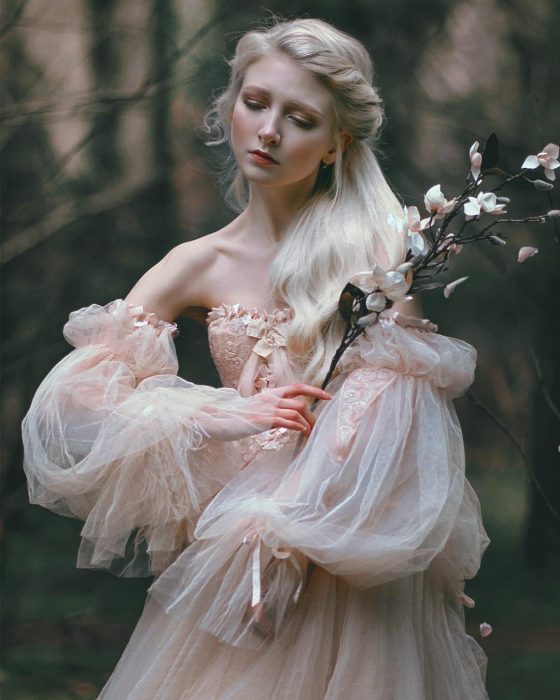 Vestidos de novia vintage confeccionados por la diseñadora Joanne Fleming, mujer rubia con vestido sin hombros, con mangas anchas sosteniendo una rama con flores