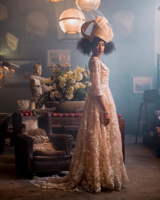 Vestidos de novia vintage confeccionados por la diseñadora Joanne Fleming, sesión de fotos antiguas con mujer morena con vestido rosa pálido largo, con mangas y encaje de flores, con un tocado de cabello con plumas, frente a un sillón de cuero café y un florero con claveles