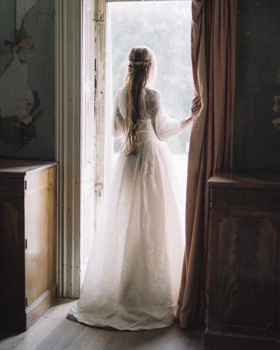 Vestidos de novia vintage confeccionados por la diseñadora Joanne Fleming, mujer de espaldas frente a una ventana, con vestido blanco largo de encaje