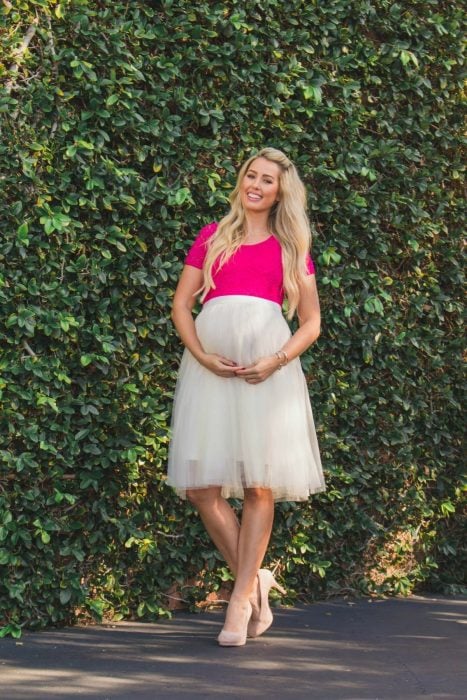 Vestidos para baby shower, chica rubia con vestido para embarazada rosa y blanco con tul, zapatillas de tacón bajo y posando frente a una pared con enredadera
