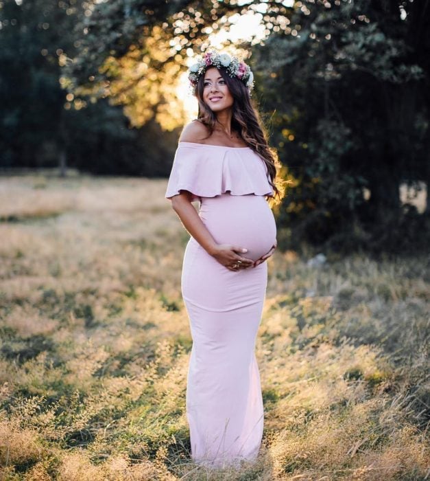 Vestidos para baby shower, mujer embarazada en sesión de fotos al aire libre con vestido rosa, largo y ceñido al cuerpo, corona de flores en la cabeza