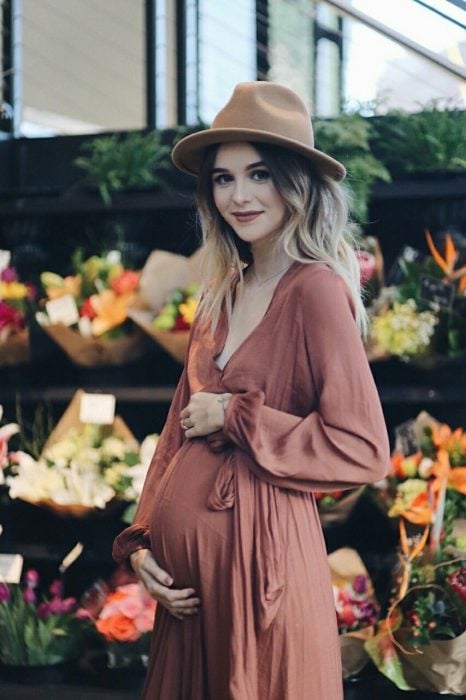 Vestidos para baby shower, chica con vestido bohemio de embarazo, color rosa y sombrero café frente a estantería con flores