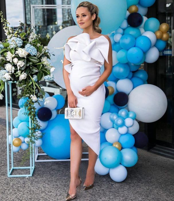 Vestidos para baby shower, mujer rubia con coleta, embarazada con vestido blanco, ceñido al cuerpo y con holanes en el pecho, tacones dorados y bolsa pequeña de mano, parada frente a globos azules