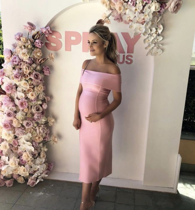 Vestidos para baby shower, mujer rubia con cabello agarrado en coleta, usando vestido para embarazada rosa, ceñido al cuerpo, de pie frente a flores roas y blancas