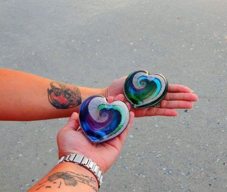 Artful ashes crea figuras de vidrio soplado con cenizas de seres queridos; mujer y hombre con tatuajes sosteneiendoadorno de cristal en forma de corazón color verde, azul y morado