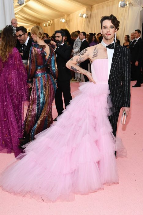 Michael Urie usando un vestido de gasa de color rosa a la mitad de su cuerpo y en la otra un traje de líneas junto con un bolso de mano