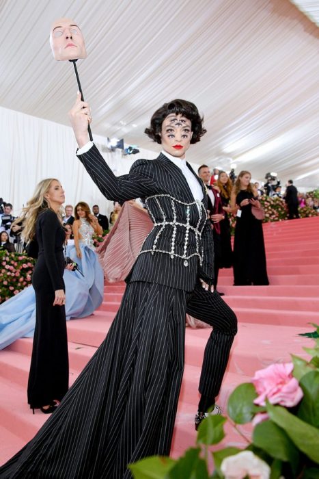 Ezra Miller usando un traje de rayas verticales con un corsaje de pedrería. Como accesorios una mascara y un maquillaje con siete ojos en su rostro 