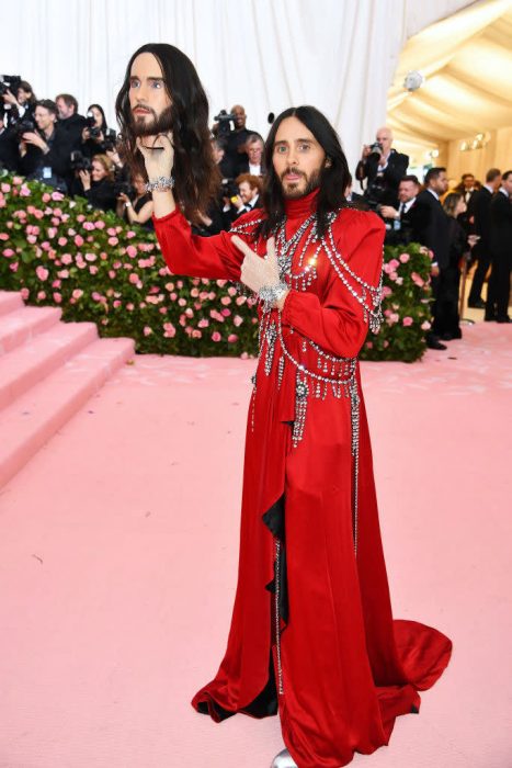 Jared Leto en la Gala MET usando un abrigo largo y rojo mientras sostiene una cabeza similar a su rostro 