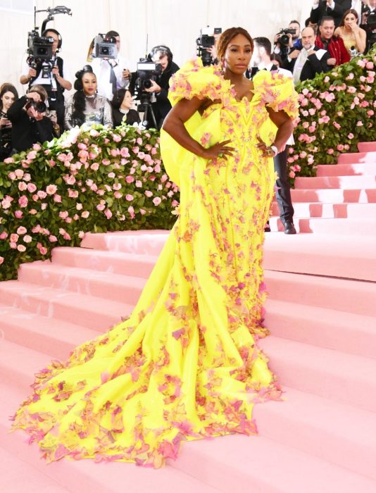 Serena Williams posando en la alfombra rosa del MET Gala. Usa un vestido amarillo con aplicaciones de flores en color rosa 