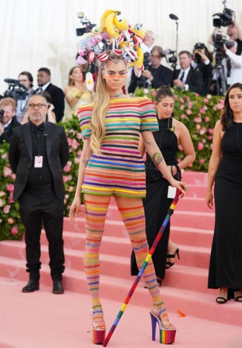 Cara Delevingne usando un vestido de lÃ­neas de diferentes colores y un sombrero con diferentes aplicaciones durante la MET Gala 2019