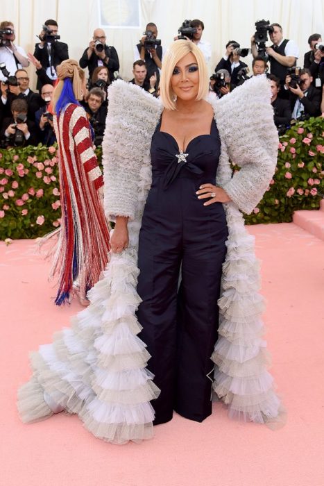 Kris Jenner en la MET Gala usando un mono de color morado con una capa de hombreras grandes en color plateado y una peluca de color rubio