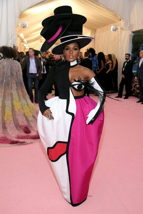 Janelle MonÃ¡e usando un traje de color rosa con blanco con estampado de un rostro y cuatro sombreros que adornan su cabeza en la Gala MET