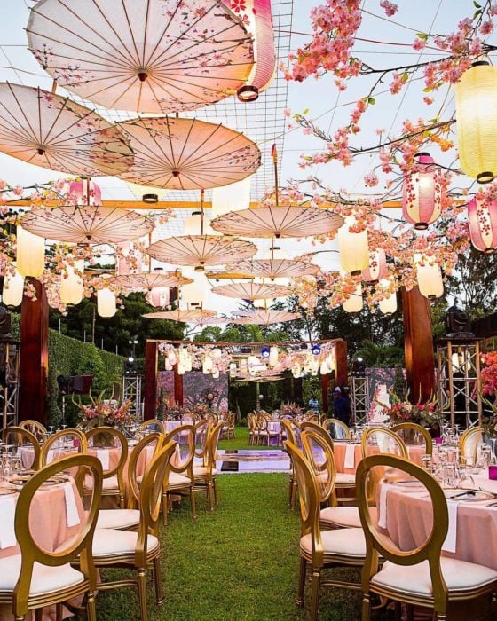 Ideas para fiesta de XV años con temática japonesa; jardín con sillas y mesas de manteles rosas con sombrillas japonesas pintadas con árboles de cerezo colgadas del techo