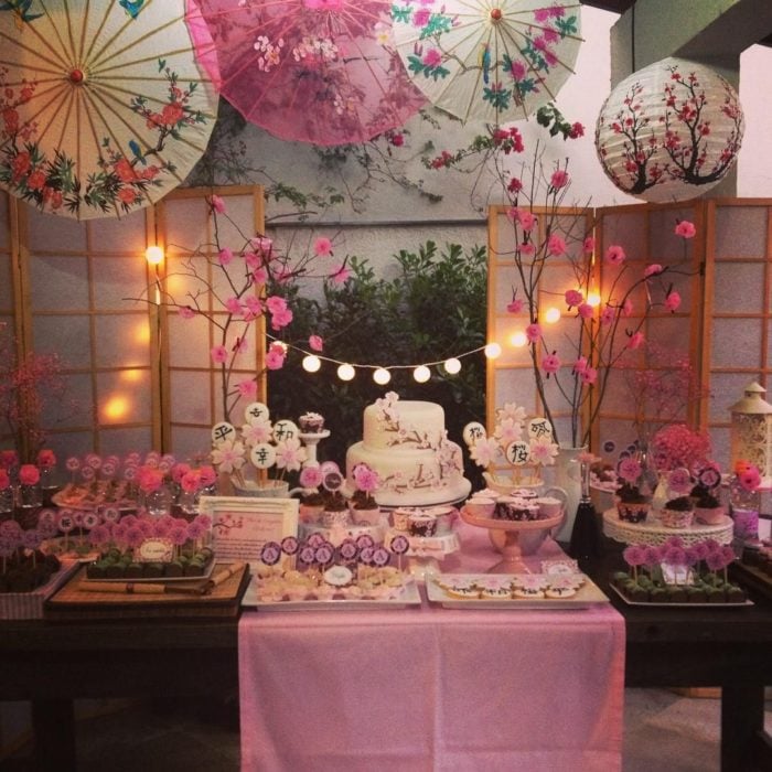 Ideas para fiesta de XV años con temática japonesa; mesa de dulces con mantel rosa, sombrillas con flores de cerezo, pastel blanco y galletas