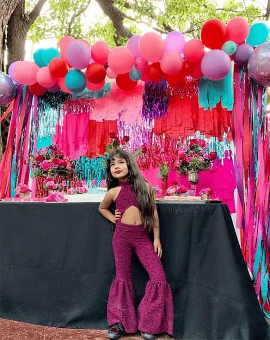 Niña parada frente a una mesa de postres decorada con globos de colores y mantel negro. Ella vistiendo un palazzo de color morado similar al de Selena 