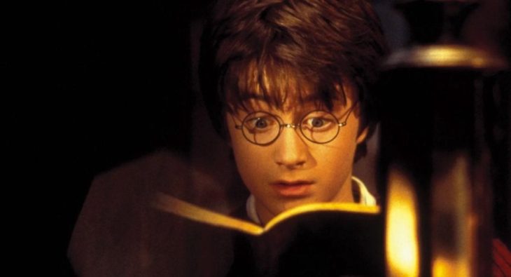 Harry Potter leyendo un libro abierto, mientras se ilumina con una vela, escena de Harry Potter y la piedra filosofal