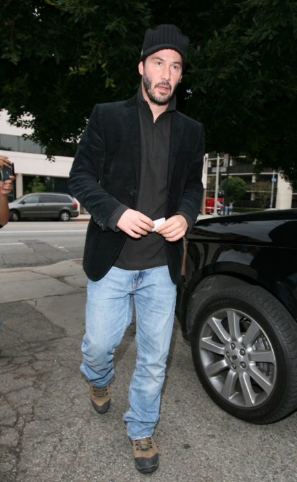 Actor Keanu Reeves, hombre caminando en la calle con gorra y saco negro, y pantalón de mezclilla