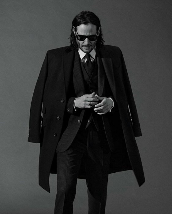 Actor Keanu Reeves, modela con traje, gabardina negra y lentes oscuros; cabello largo y barba para Saint Laurent