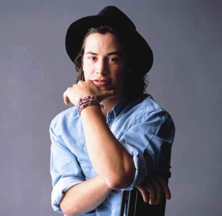 Actor Keanu Reeves; joven con camisa azul, sombrero negro y cabello largo