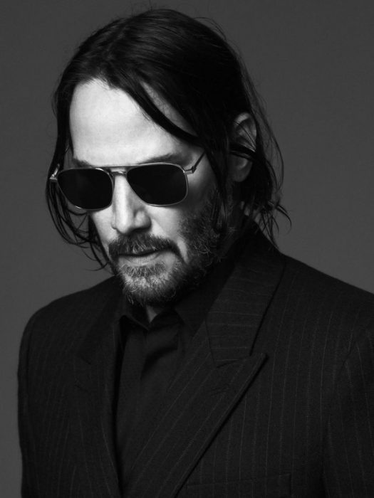 Keanu Reeves con cabello largo y gafas oscuras mirando hacia abajo mientras modela para la nueva campaña de Saint Laurent