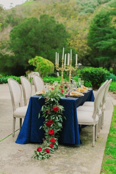 Ideas para quinceañera estilo La Bella y la Bestia de Disney; fiesta al aire libre, mesa con mantel azul y sillas blancas, adornada con flores y candelabros 