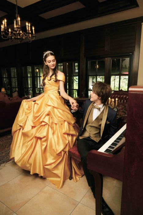 Ideas para quinceañera estilo La Bella y la Bestia de Disney; sesión de fotos de pareja con temática de princesas; mujer con vestido amarillo y tiara, hombre con traje azul y dorado sentado en un piano
