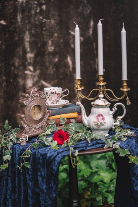 Ideas para quinceañera estilo La Bella y la Bestia de Disney; objetos vintage, reloj, taza, tetera y candelabro, con libros viejos y mantel de terciopelo azul