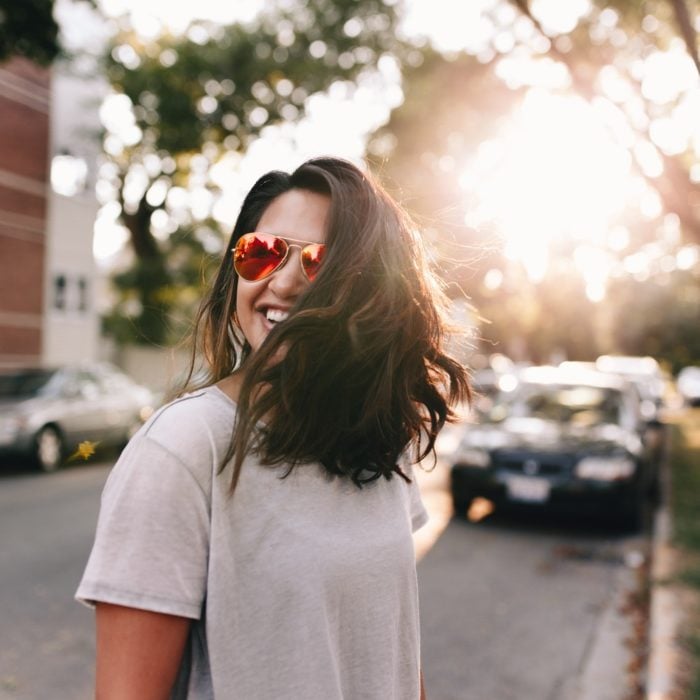 Mujer con gafas rojas a mitad de la avenida durante el atardecer sonriendo y cubriendo su rostro con su propio cabello