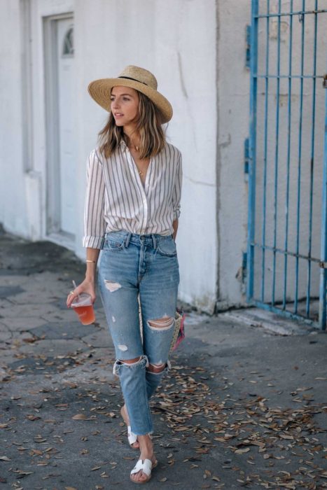 Looks de primavera; chica caminando con sombrero de mimbre, blusa de rayas, pantalón de mezclilla recto y desgastado y sandalias blancas de piso; con bebida en la mano