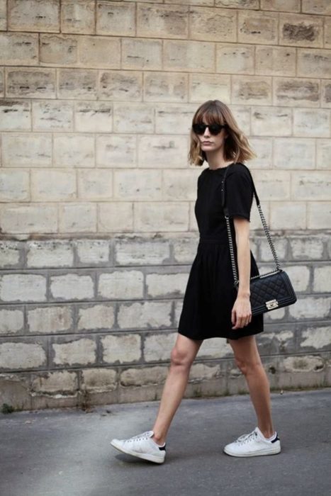 Looks de primavera; joven de cabello castaño hasta los hombros, con vestido negro sencillo, con mangas, bolsa de mano negra con cadena y tenis blancos