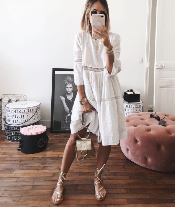 Looks de primavera; mujer de cabello corto y rubio, con vestido blanco de algodón, bolsa de mano y huaraches dorados, tomándose selfie en su cuarto frente al espejo