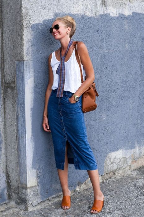 Looks de primavera; mujer rubia sonriendo recargada en la pared, usando blusa básica sin mangas, falda larga de mezclilla, sandalias anaranjadas y corbata