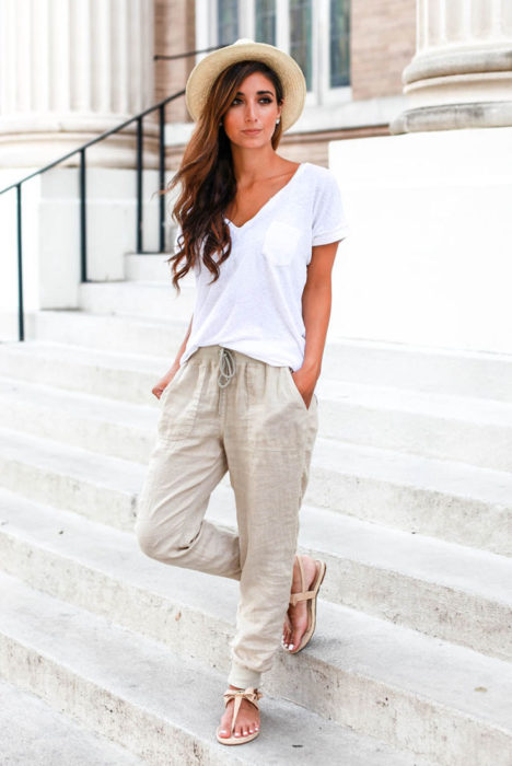 Looks de primavera; mujer de cabello castaño, ondulado y largo, con sombrero de mimbre, blusa blanca básica y pantalón beige holgado de lino delgado y sandalias de piso