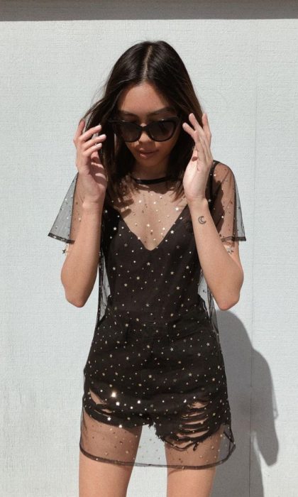 Looks de primavera; muchacha con vestido negro con transparencia de gasa y estrellas doradas; tatuaje de luna en la muñeca