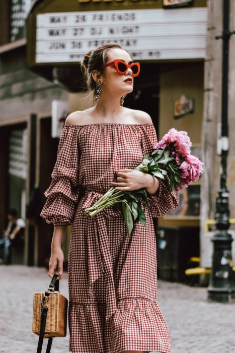 Looks de primavera; chica con vestido estilo campesino de cuadros rojos y blancos, con lentes de marco grueso y rojo, con bolsa de mano de mimbre, sosteniendo un ramo de flores rosas