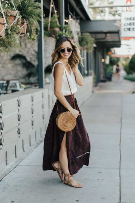 Looks de primavera; mujer con blusa de algodón blanca, falda larga color vino floreada, sandalias de piso y bolsa de mano de mimbre; cabello rubio ombré