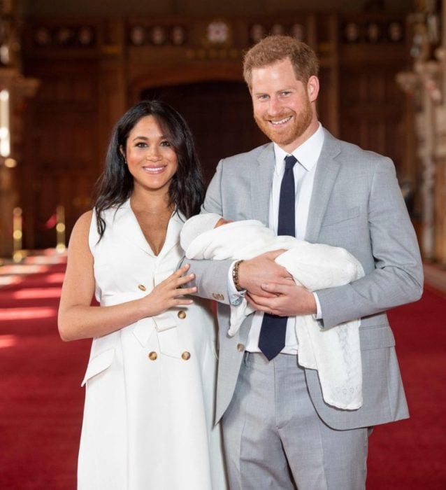 Meghan Markle y el príncipe Harry caminando sobre una alfombra roja y cargando a su primogénito en brazos