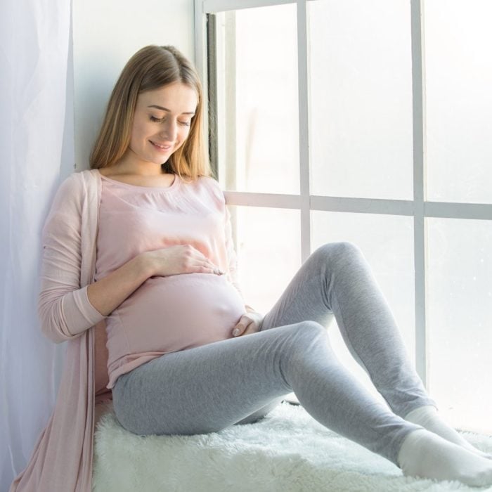 Mujer joven embarazada tocándose el vientre mientras está sentada junto a la ventana