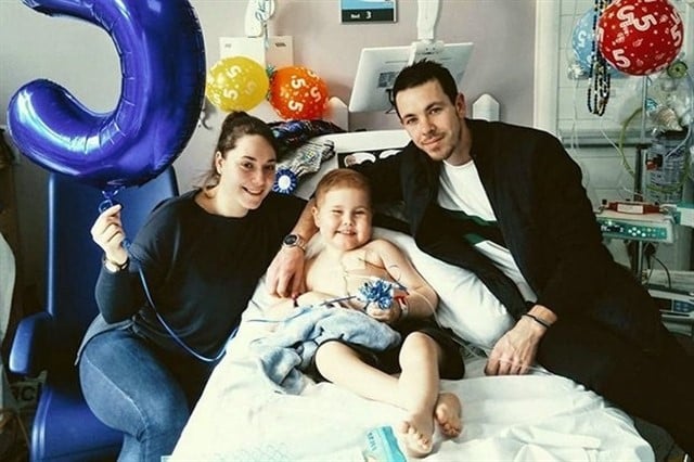 Oscar Saxelby-Lee, niño con cáncer, sentado en una cama de hospital junto a sus padres, todos sonriendo para una fotografía mientras sostienen globos de helio