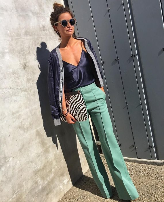 Chica parada mientras posa para una sesión de fotos mientras usa un pantalón de vestir verde, blusa de seda azul, bolso animal print y chaqueta azul 