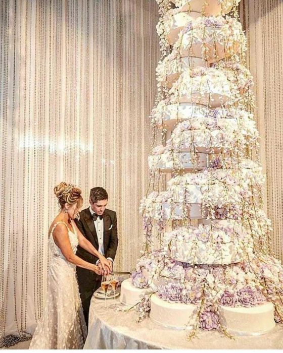 Pareja de novios el día de su boda parados frente a un enorme pastel decorado con flores moradas 