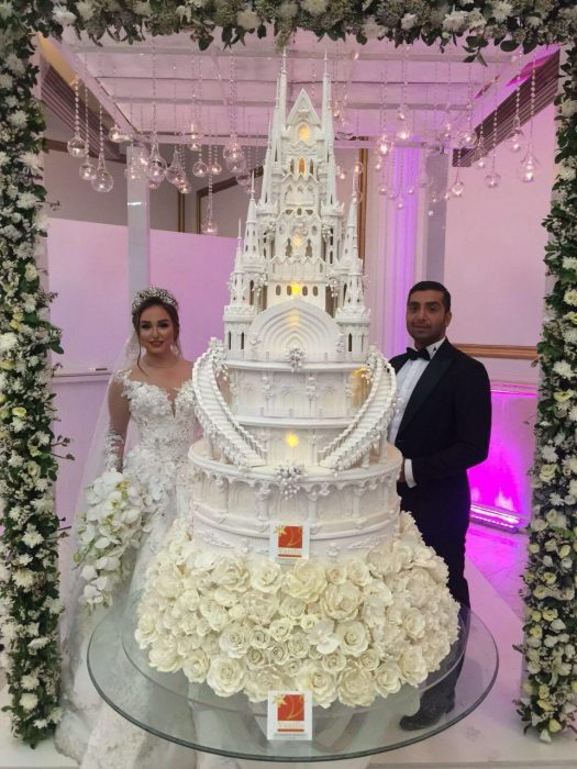 Pareja de novios el día de su boda parado a un lado de su enorme pastel de bodas con diseño de un castillo 