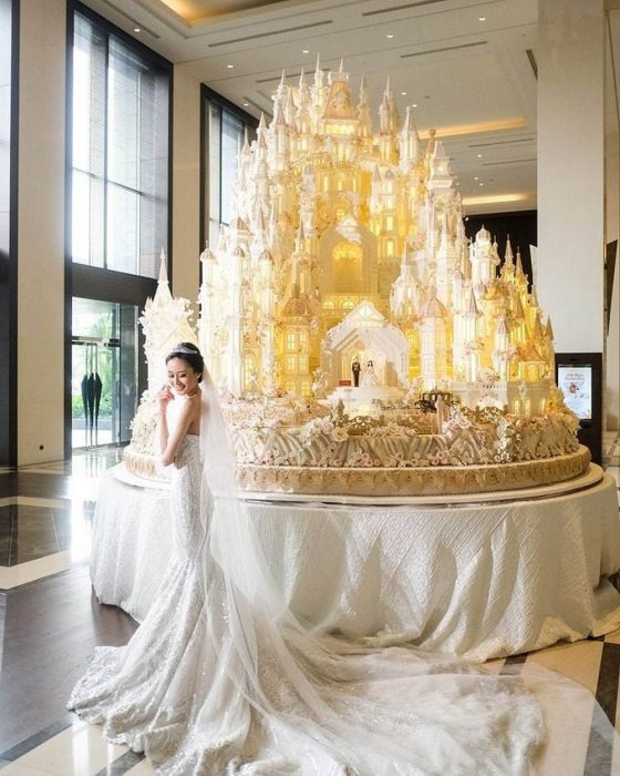 Novia el día de su boda parada frente a un enorme pastel de bodas en forma de castillo 