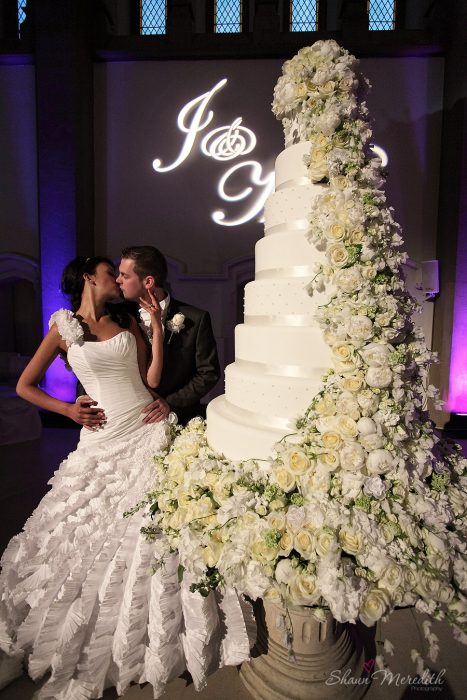 Pareja de novios parados frente a su enorme pastel de bodas decorado con flores rosas mientras se dan un beso 