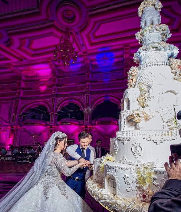 Pareja de novios el día de su boda partiendo un pastel de boda con 8 pisos, de color blanco, con flores de azúcar y decoraciones con betún 