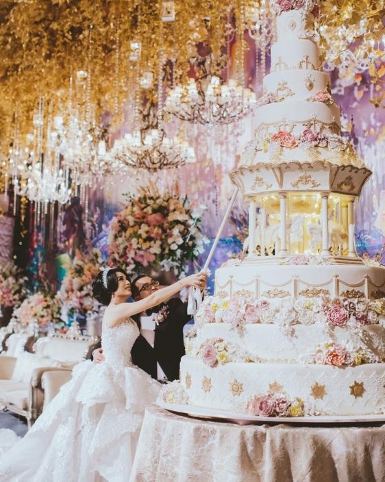 Pareja de novios el día de su boda cortando un enorme pastel de 9 pisos de color blanco y decorado con rosas 